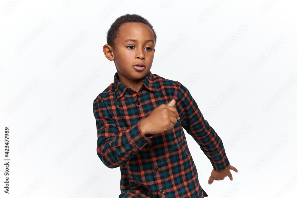 Action shot of adorable active dark skinned little boy wearing casual checkered shirt moving arms as if walking, dancing or doing physical exercises, having confident facial expression
