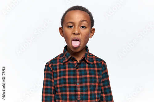 Portrait of nasty mischievous little black boy grimacing, making disgusting facial expression, sticking out tongue, acting naughty, teasing you, misbehaving. Childhood, upbringing and bad behavior