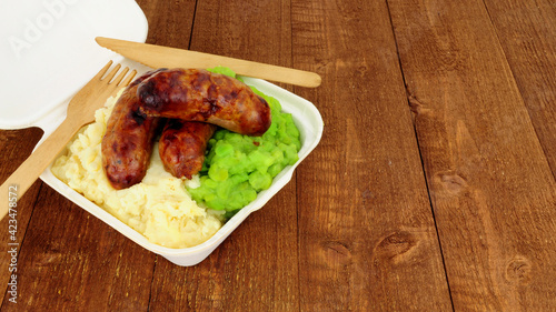 Sausages and mashed potato with mushy peas meal in a take away box on a wooden background