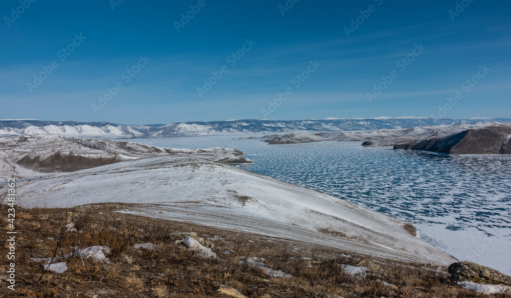 Panorama of the frozen lake. The icy surface is covered with snow, like lace. Around are snow-capped hills. Dry grass in the foreground. Sunny winter day, blue sky. Baikal