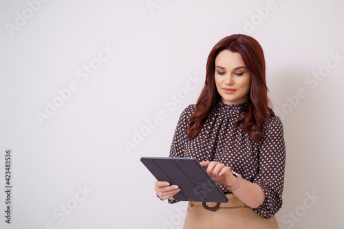 Beautiful modern woman having tablet in hands, leaning on a white wall