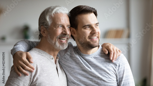 Fotografia Smiling young Caucasian man and older father hug look in window distance dreaming imagining together