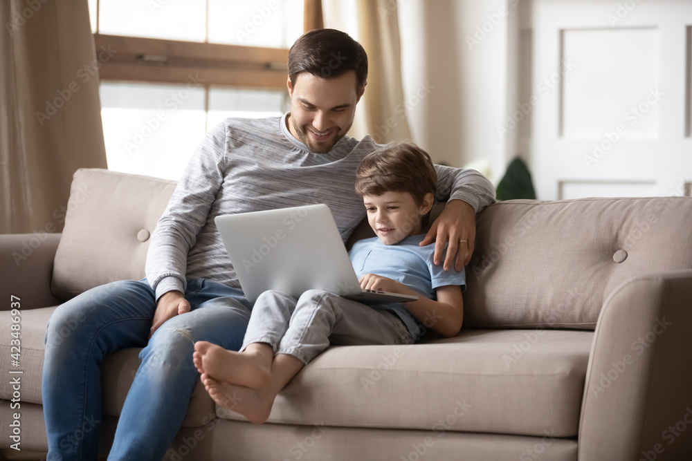 Loving happy young Caucasian father and little 7s son relax on sofa at home use modern laptop studying online. Smiling dad and small kid child look at computer screen talking on webcam call together.