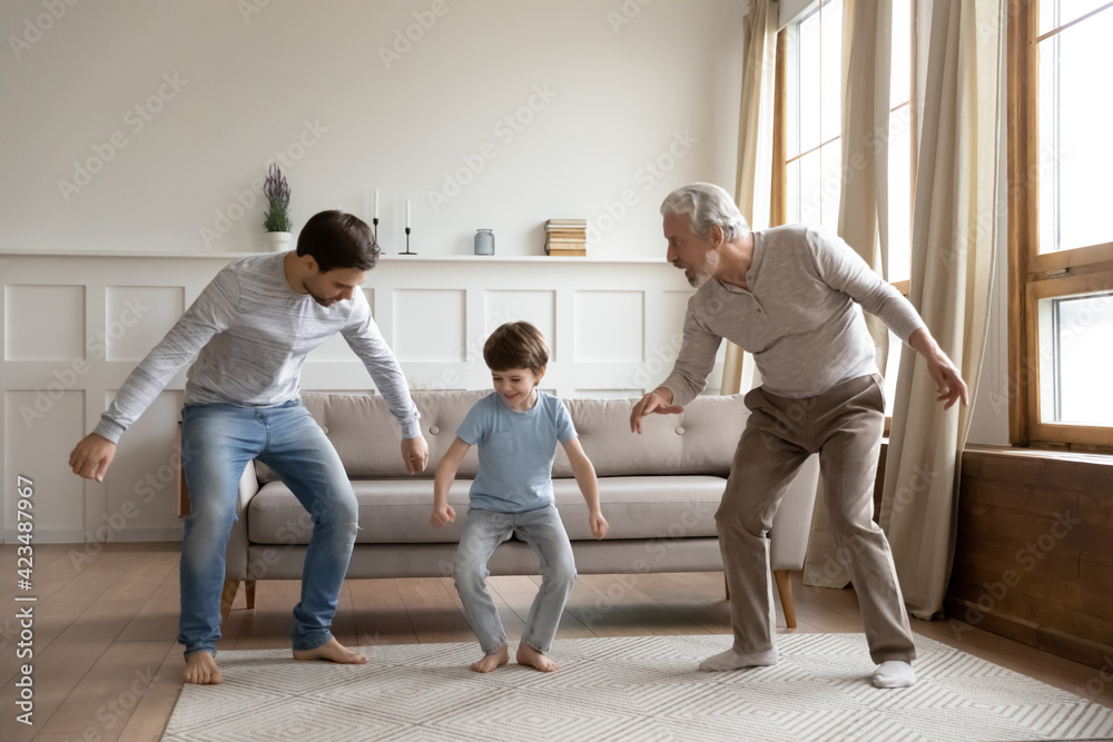 Fototapeta premium Happy three generations of Caucasian men have fun dancing together in cozy living room on weekend. Overjoyed little boy child with young father and old grandfather involved in funny game at home.