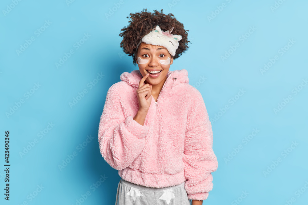 Horizontal shot of beautiful cheerful Afro American woman has positive face expression dressed in soft pajama hears good news in morning isolated over blue background. Awakening and rest concept Horizontal shot of beautiful cheerful Afro American woman has positive face expression dressed in soft pajama hears good news in morning isolated over blue background. Awakening and rest concept