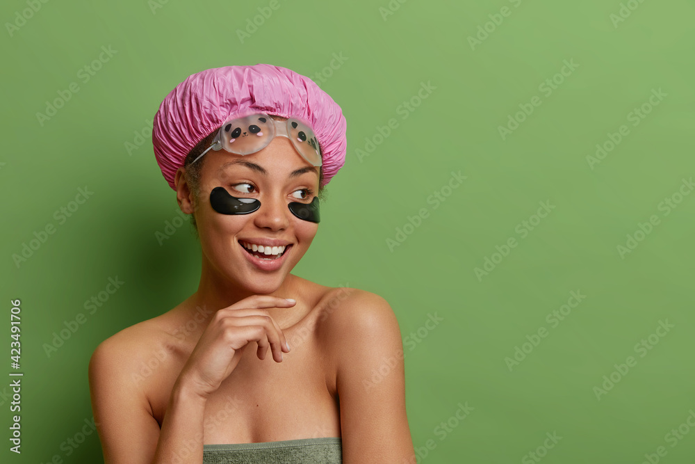 Studio shot of lovely cheerful woman keeps hand under chin touches gently jawline wears waterproof hat bath towel around naked body isolated over green background copy space aside for your promotion Studio shot of lovely cheerful woman keeps hand under chin touches gently jawline wears waterproof hat bath towel around naked body isolated over green background copy space aside for your promotion
