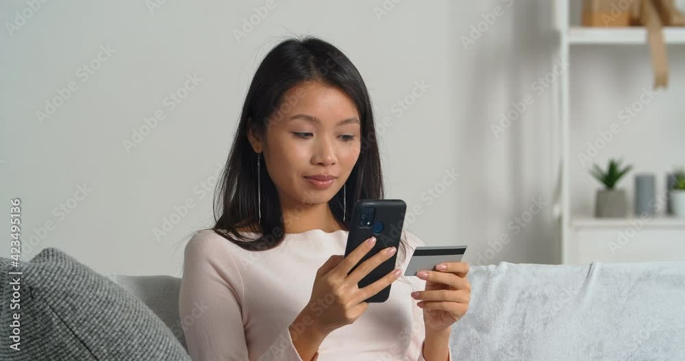 Asian woman sitting at home couch holds credit card in her hand and mobile phone enters secret bank account number confirming transaction onInternet shop online book plane tickets orders goods in net Asian woman sitting at home couch holds credit card in her hand and mobile phone enters secret bank account number confirming transaction onInternet shop online book plane tickets orders goods in net