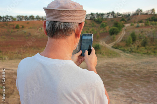 Older man in a white T-shirt and sun hat making picture of summer landscape with field, slopes and sky on smartphone. Back view. 