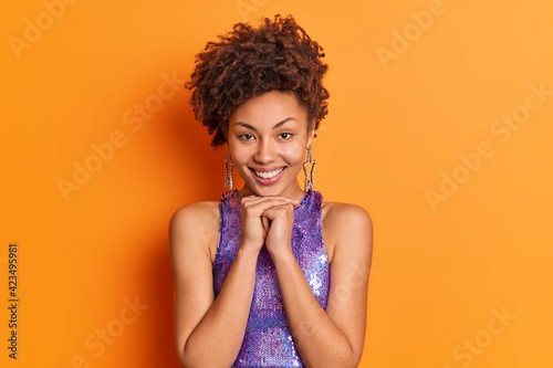 Pretty lovely fashionable woman with curly hair keeps hands under chin smiles positively dressed in stylish clothes expresses positive emotions isolated over vivid orange background. Emotions concept