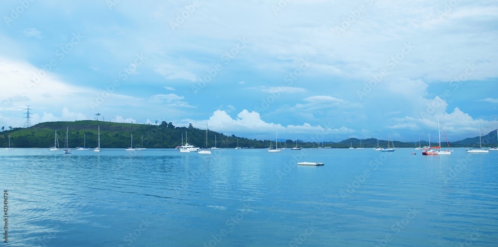 Panoramic view of sea bay. Land with green hills on the horizon. Blue sky with clouds. Calm water. Sailing yachts in the harbor. Wide frame, seascape, tropical resort. Thailand, Phuket island, boats