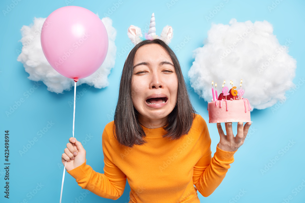 Bad holiday concept. Sad stressful Asian woman cries from despair holds inflated balloon and tasty dessert has unhappy mood on birthday upset none came to congratulate her has mournful look. - obrazy, fototapety, plakaty 