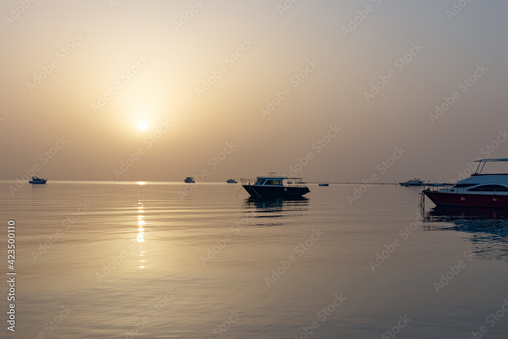 Dawn on the Red Sea in Egypt, Makadi Bay, calm sea, silhouettes of boats, golden disc of the sun on the horizon, reflection of sunlight on the water surface
