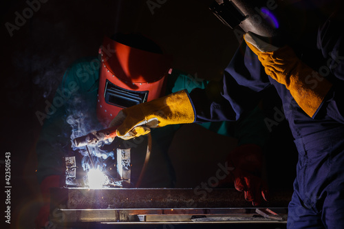 a factory worker wearing a green mechanic coveralls and safety helmet welding metalwork at night time in a factory while his colleague looking at a spark from welding