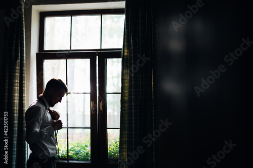Elegant bearded groom standing by the window. Businessman in a hotel room. The man at the window in the room. Good life and easy relaxing business person lifestyle after hotel work.