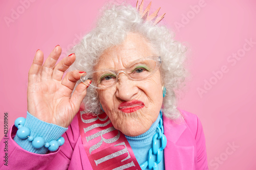 Portrait of grey haired wrinkled woman pouts lips looks attentively at camera keeps hand on rim of glasses dressed in fashionable clothes isolated over pink background waits for party to start