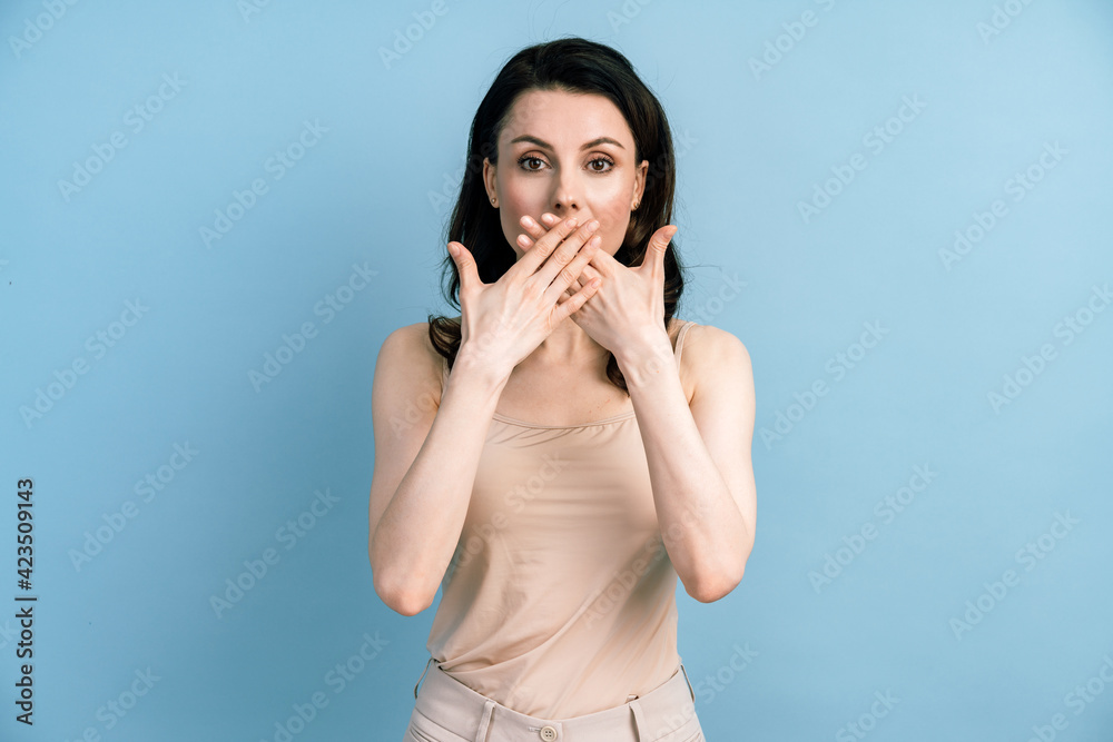 Surprised worried brunette woman in t-shirt closing her mouth and looking at camera on blue background. Cute woman posing isolated on blue background. Surprised worried brunette woman in t-shirt closing her mouth and looking at camera on blue background. Cute woman posing isolated on blue background.