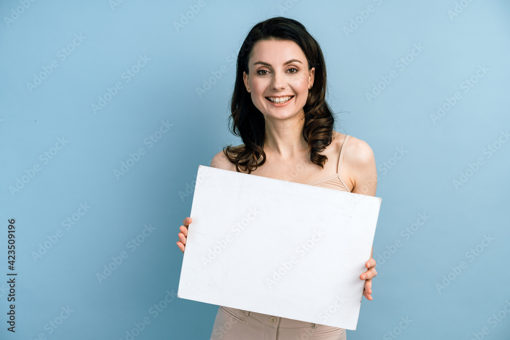Beautiful brunette girl with holds a white sheet of paper on a blue background. Place for text, banner, surface for inscriptions and advertising. Beautiful brunette girl with holds a white sheet of paper on a blue background. Place for text, banner, surface for inscriptions and advertising.