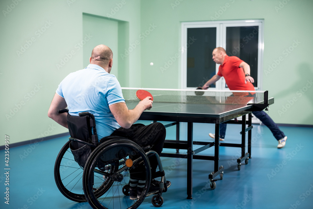 Adult disabled man in a wheelchair play at table tennis with his coach