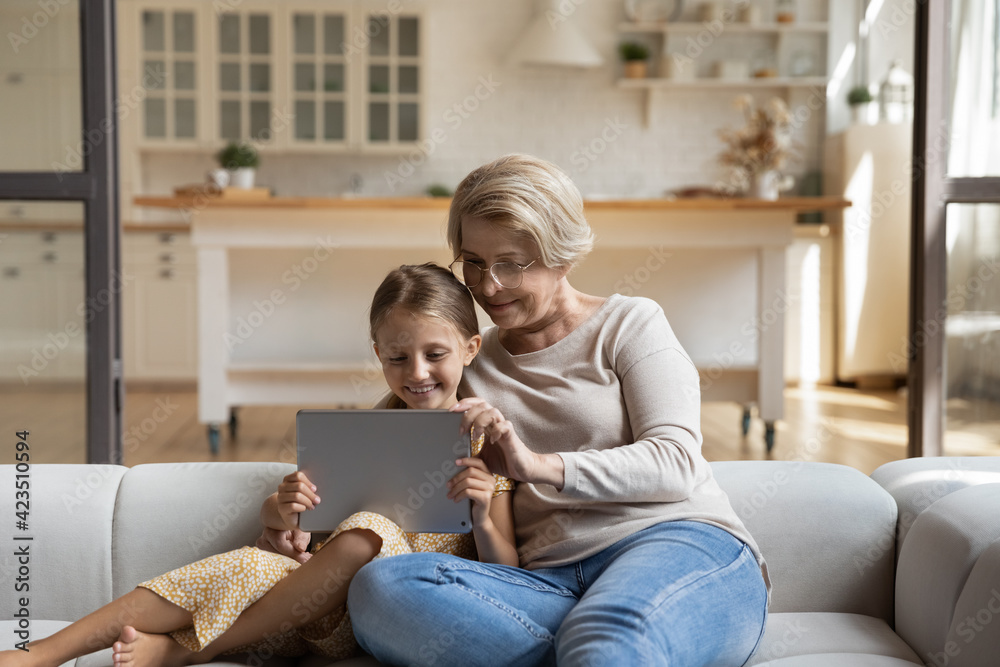 Happy loving elderly Caucasian grandmother and 7s granddaughter relax on sofa in living room using tablet together. Smiling mature granny and small girl child have fun with pad. Technology concept.