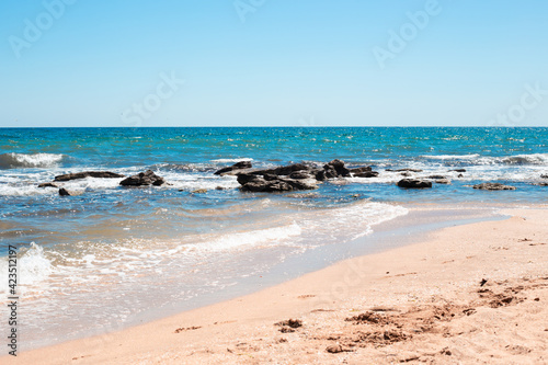 Seashore with blue water beating against stones and a sandy beach. Sea sunny summer landscape