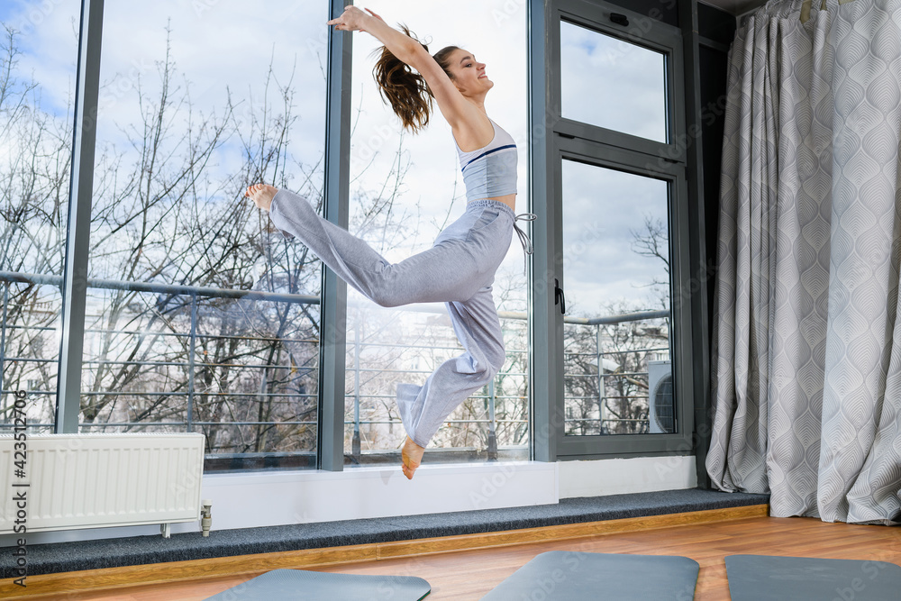 Young sport woman jumping high barefoot at light studio hall, wearing cozy grey pants and top, healthy active lifestyle