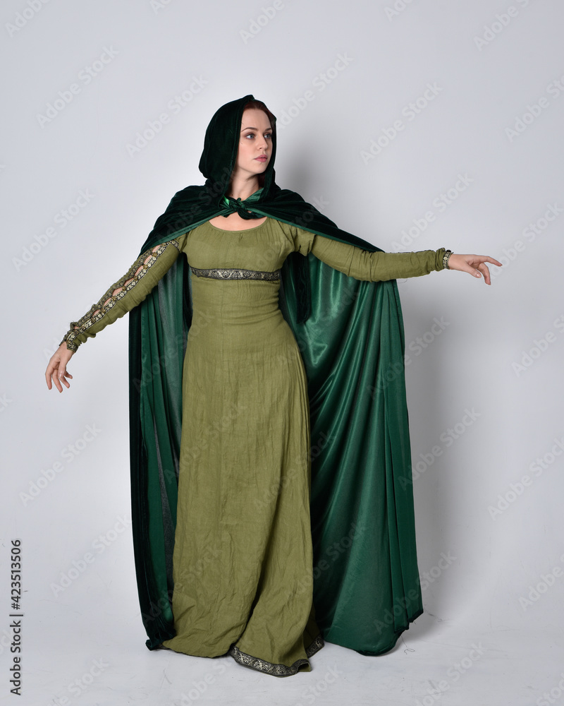 Naklejka premium full length portrait of red haired girl wearing celtic, green medieval gown with fantasy velvet cloak. Standing pose isolated against a studio background.