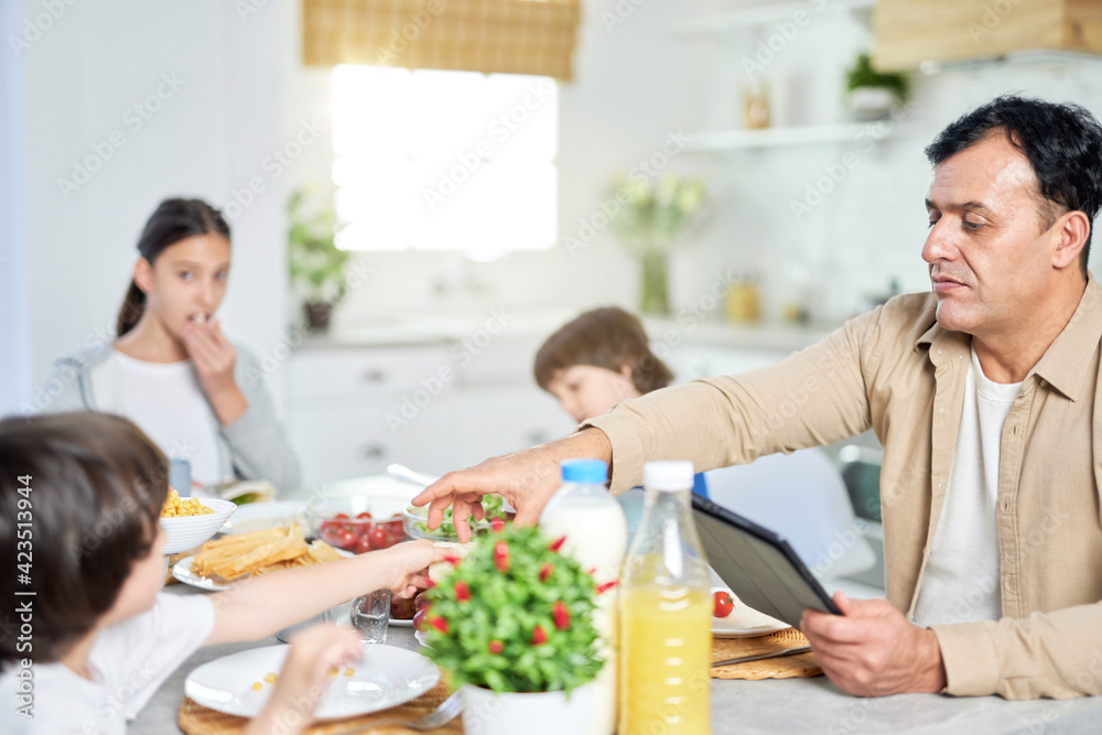 Middle aged father enjoying meal together with his family, using digital tablet while sitting at the table in kitchen at home Middle aged father enjoying meal together with his family, using digital tablet while sitting at the table in kitchen at home