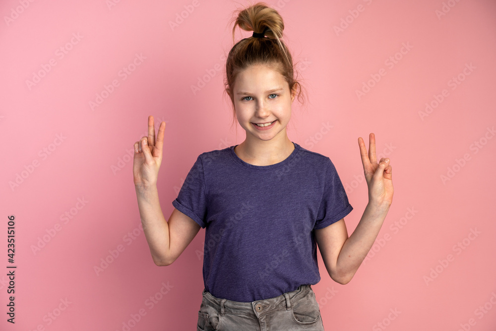 Cheerful child gestures, shows a gesture of peace, victory on the background of a pink wall. Teen girl on a copy space background.