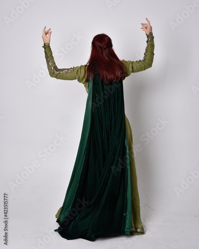 full length portrait of red haired girl wearing celtic  green medieval gown with fantasy velvet cloak. Standing pose isolated against a studio background.