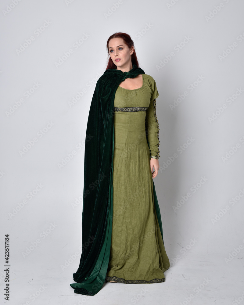 full length portrait of red haired girl wearing celtic, green medieval gown with fantasy velvet cloak. Standing pose isolated against a studio background. full length portrait of red haired girl wearing celtic, green medieval gown with fantasy velvet cloak. Standing pose isolated against a studio background.