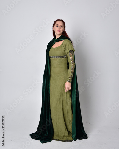 full length portrait of red haired girl wearing celtic, green medieval gown with fantasy velvet cloak. Standing pose isolated against a studio background.