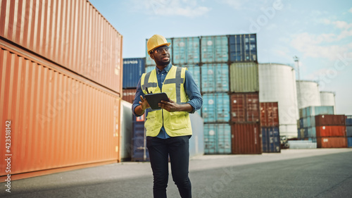 Smiling Handsome African American Black Industrial Engineer in Yellow Hard Hat and Safety Vest Working on Tablet Computer. Foreman or Supervisor in Container Terminal.