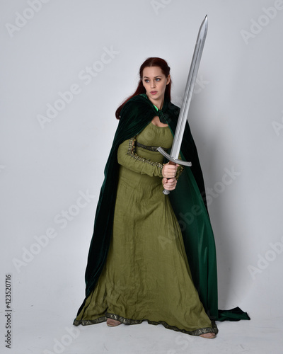 full length portrait of red haired girl wearing celtic, green medieval gown with fantasy velvet cloak. Standing pose isolated against a studio background.