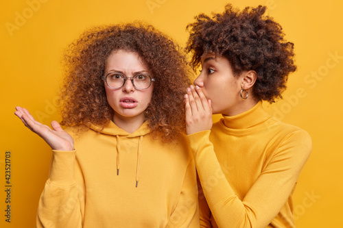 Surprised Afro American woman whispers secret to friend tells secretly something on ear share gossips dressed casually isolated over yellow background. Puzzled curly European female hears rumor