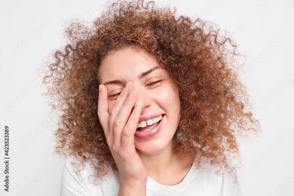 Headshot of beautiful sincere European woman with curly bushy hair has fun laughs out covers face with palms smiles broadly expresses happiness isolated over white background. Positive emotions Headshot of beautiful sincere European woman with curly bushy hair has fun laughs out covers face with palms smiles broadly expresses happiness isolated over white background. Positive emotions