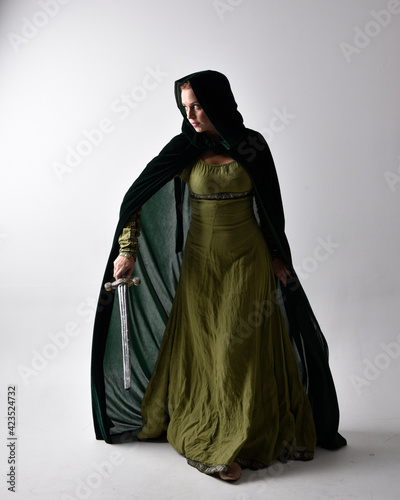 full length portrait of red haired girl wearing celtic, green medieval gown and velvet cloak with shadowy backlighting. Standing pose isolated against a studio background.