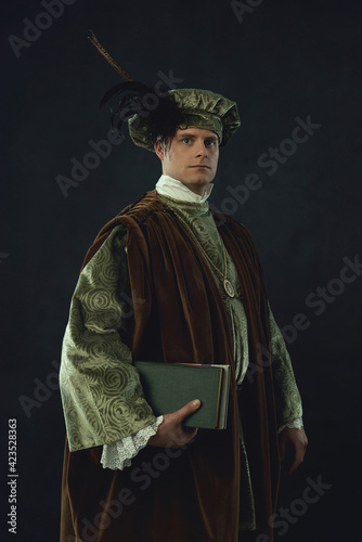 Obraz na płótnie Renaissance man holding a book stands in front of a dark wall. w salonie