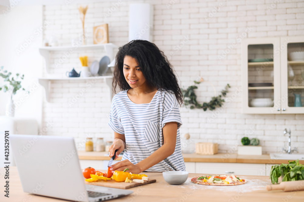Happy beautiful biracial woman enjoying cooking and filming it, looking for a recipe on the laptop in the modern kitchen, food blogger concept, healthy lifestyle, online learning cooking class concept