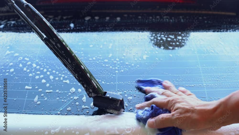 Man wearing an orange shirt is washing a white car with blue cloth, car