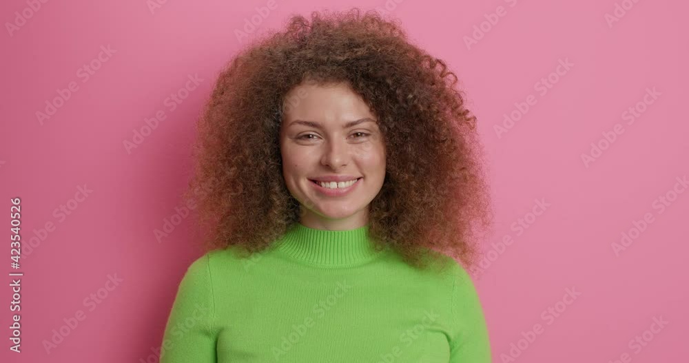 Pretty curly haired woman smiles gentlly happy to hear something pleasant shows white teeth dressed in casual green jumper isolated over pink bbackground has friendly expression. Emotions concept Pretty curly haired woman smiles gentlly happy to hear something pleasant shows white teeth dressed in casual green jumper isolated over pink bbackground has friendly expression. Emotions concept