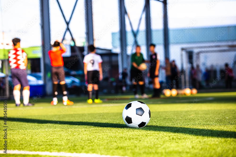 Obraz premium Soccer ball on green artificial turf with blurry coach and soccer team are training. Blurry kid soccer players training in soccer academy.