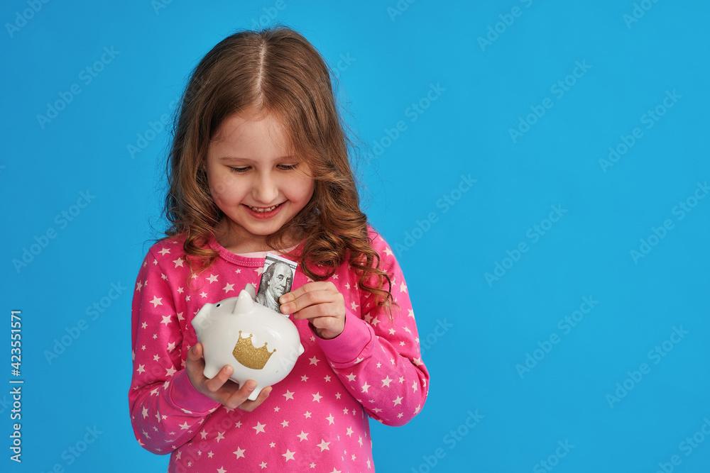 Happy savings. A little cute girl with a piggy bank on a blue background. The child smiles happily and puts money in the piggy bank The concept of saving money for a dream.