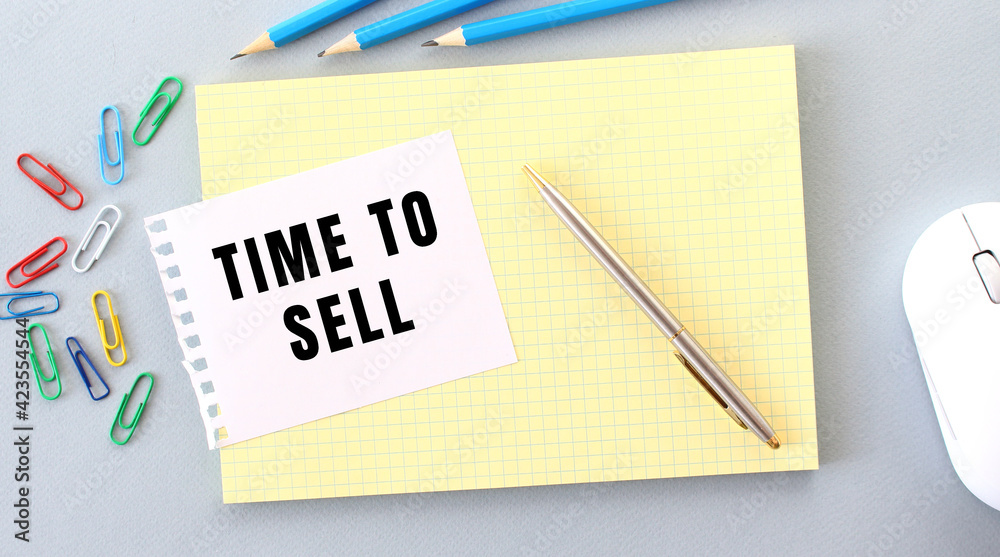 TIME TO SELL is written on a piece of paper that lies on a notebook next to office supplies. TIME TO SELL is written on a piece of paper that lies on a notebook next to office supplies.