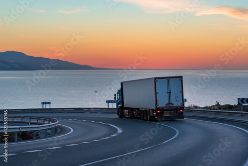 Truck with refrigerated semi-trailer on a mountain road with the sea and sunrise on the horizon.