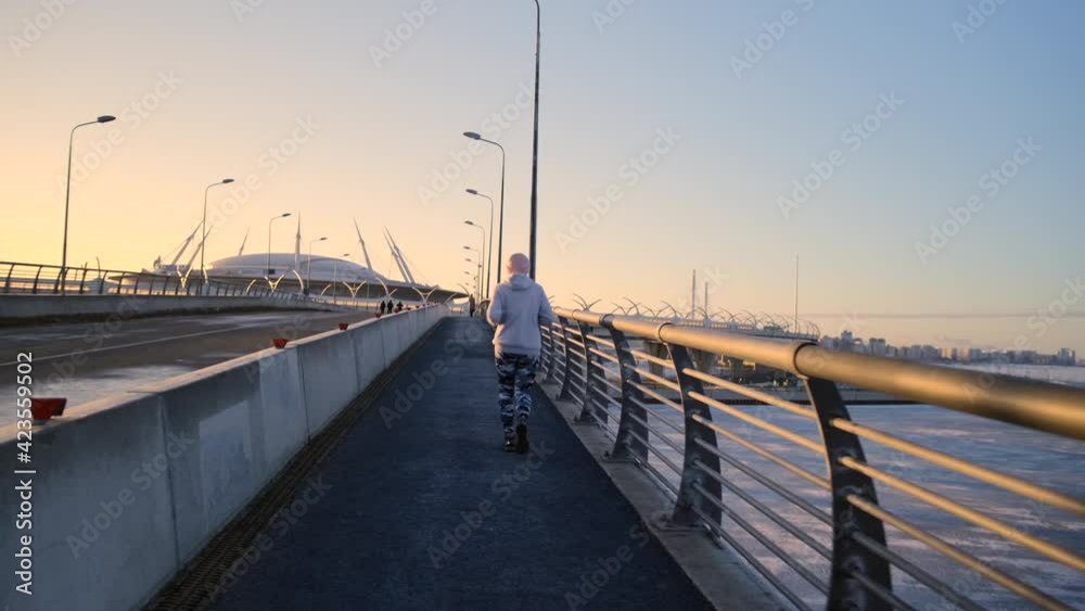 Back view of a girl running across the bridge in tight camouflage leggings, a white sweater and a pink hat. Morning jog overlooking the frozen river and stadium. Back view of a girl running across the bridge in tight camouflage leggings, a white sweater and a pink hat. Morning jog overlooking the frozen river and stadium.