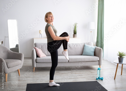 Happy senior woman doing flexibility exercises, stretching her leg on yoga mat at home, full length