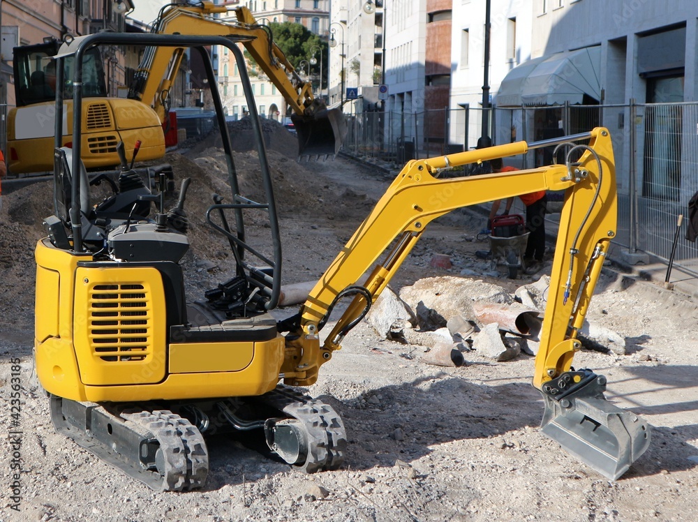A mini excavator with a large excavator behind it at work in the renovation of the road surface in an urban street.