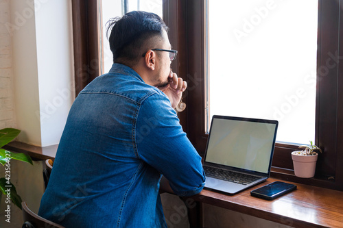 Close up of a asian man wears blue jeans jacket working, thinking and using laptop with white mockup screen in a coffee shop