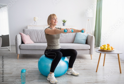Strong mature woman working out with dumbbells on fitness ball at home, blank space photo