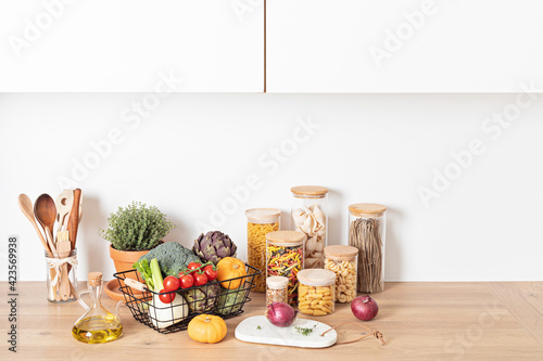 Assortment of pasta in glass jars, olive oil, vegetables and kitchen utensils on wooden table. Traditional italian ingredients, healthy balanced food, sustainable lifestyle, zero waste concept.